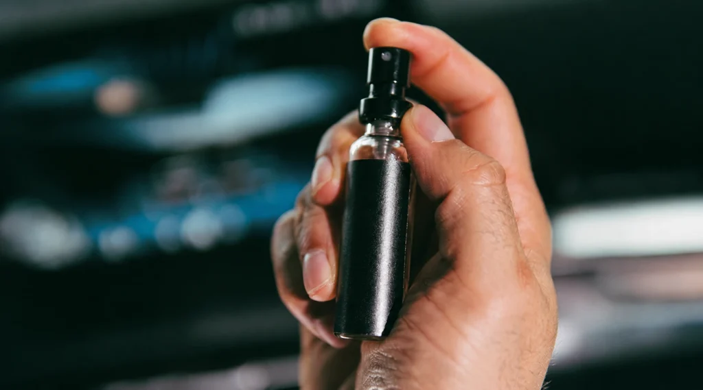 Foto de mão segurando frasco de perfume veicular dentro de carro.