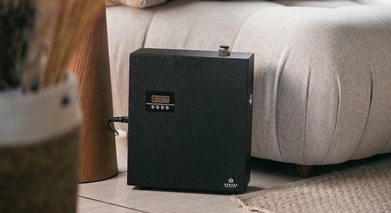 Dispositivo aromatizador de ambientes preto da Senses colocado no chão ao lado de um sofá bege em ambiente interno, com parte de vaso decorativo e tapete visível.