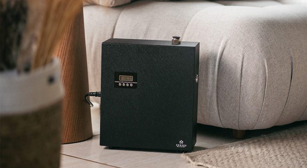 Dispositivo aromatizador de ambientes preto da Senses colocado no chão ao lado de um sofá bege em ambiente interno, com parte de vaso decorativo e tapete visível.