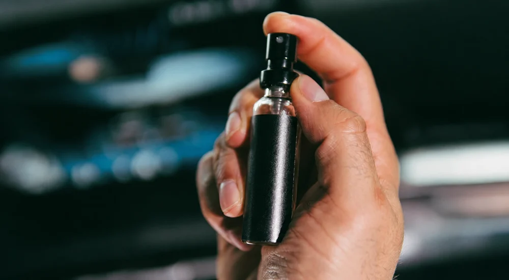 Foto de mão segurando frasco de perfume veicular dentro de carro.